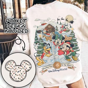 May include: Cream-colored sweatshirt featuring a festive Disney design. The graphic includes Mickey, Minnie, Donald, and Pluto in winter attire, with a snowy cabin and the text "Walt Disney World". A pocket design with Mickey Mouse ears and string lights is also included.