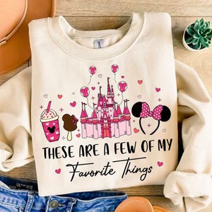 May include: A cream-colored sweatshirt with a pink and white graphic design. The design features a cartoon image of Cinderella's castle, Mickey Mouse ears, a Mickey Mouse ice cream bar, and a Mickey Mouse milkshake. The text "These are a few of my favorite things" is written in black below the graphic.