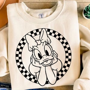 K&ouml;nnte beinhalten: Cremefarbenes Sweatshirt mit einer schwarz-wei&szlig;en Grafik von Daisy Duck in einem karierten Kreis. Die Zeichentrickfigur st&uuml;tzt ihr Kinn auf ihre H&auml;nde. Im Hintergrund ist eine kleine Sukkulentenpflanze zu sehen.