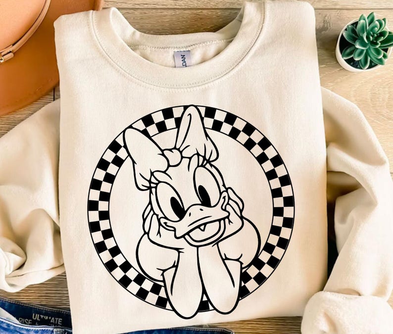K&ouml;nnte beinhalten: Cremefarbenes Sweatshirt mit einer schwarz-wei&szlig;en Grafik von Daisy Duck in einem karierten Kreis. Die Zeichentrickfigur st&uuml;tzt ihr Kinn auf ihre H&auml;nde. Im Hintergrund ist eine kleine Sukkulentenpflanze zu sehen.