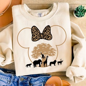 May include: A beige sweatshirt with a brown and black leopard print bow on a brown Mickey Mouse silhouette. The silhouette includes a brown tree with a monkey, an elephant, a giraffe, a lion, and a deer.