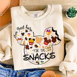 May include: A white sweatshirt with a black Mickey Mouse head design. The Mickey Mouse head is filled with various Disney snacks and the text "I'm just here for the snacks".