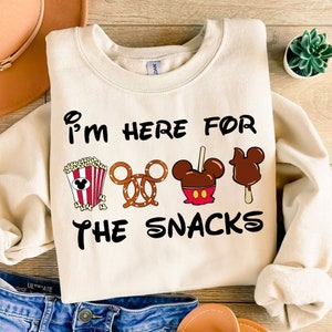 May include: A cream-coloured sweatshirt with the text "I'm here for the snacks" printed in black. The text is surrounded by cartoon images of popcorn, a pretzel, a chocolate-covered apple, and a Mickey Mouse-shaped ice cream bar.