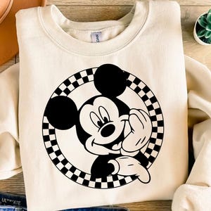 K&ouml;nnte beinhalten: Cremefarbenes Sweatshirt mit einem schwarz-wei&szlig;en Mickey Mouse-Motiv. Die Grafik zeigt Mickey Mouse, die in einem karierten Kreis l&auml;chelt. Das Sweatshirt hat einen Rundhalsausschnitt und lange &Auml;rmel.