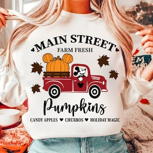 Op de afbeelding: Witte sweatshirt met een rode vrachtwagenafbeelding met pompoenen met Mickey Mouse oren. De tekst luidt "Main Street Farm Fresh Pumpkins" met de toevoegingen "Candy Apples, Churros, Holiday Magic".