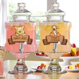 May include: Two glass beverage dispensers with spouts, one pink with Winnie the Pooh and the text "Pooh's Juice", the other yellow with Eeyore and the text "Eeyore's Lemonade".