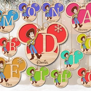 Op de afbeelding: Houten ornamenten in de vorm van Mickey Mouse-oren met een personage uit de Toy Story-film in het midden. Elk ornament heeft een andere letter en naam erop gedrukt. De namen zijn Mason, Oliver, Nathan, Andrew, Brian, Daniel, Lucas, Grace, Fiona, Isaac, Harper en Calde.