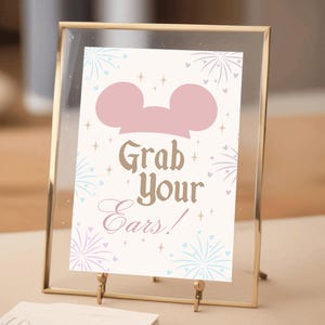 Puede incluir: Un cartel con marco dorado, fondo blanco y orejas de Mickey Mouse rosas. El cartel dice "Grab Your Ears!" con un diseño de fuegos artificiales rosa y azul.