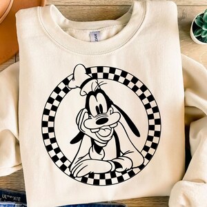 K&ouml;nnte beinhalten: Cremefarbenes Sweatshirt mit einer schwarz-wei&szlig;en Grafik von Goofy von Disney. Die Grafik befindet sich in einem karierten Kreis. Das Sweatshirt hat einen Rundhalsausschnitt und lange &Auml;rmel. Eine kleine Sukkulentenpflanze ist sichtbar.