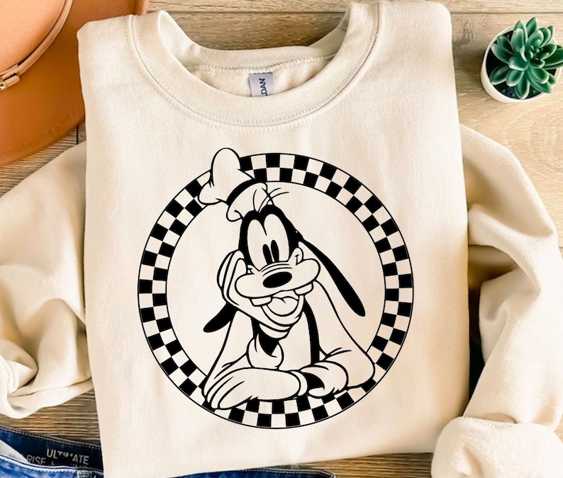 K&ouml;nnte beinhalten: Cremefarbenes Sweatshirt mit einer schwarz-wei&szlig;en Grafik von Goofy von Disney. Die Grafik befindet sich in einem karierten Kreis. Das Sweatshirt hat einen Rundhalsausschnitt und lange &Auml;rmel. Eine kleine Sukkulentenpflanze ist sichtbar.