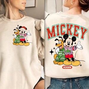 May include: A beige sweatshirt with a Christmas-themed Mickey Mouse and Pluto graphic on the front and the text "MICKEY SEASON" on the back.