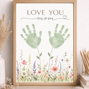 Könnte beinhalten: Ein gerahmter Druck mit den Worten "LOVE YOU mom" und zwei grünen Handabdrücken. Die Handabdrücke befinden sich über einer Aquarellillustration von bunten Wildblumen. Der Rahmen ist aus hellem Holz.