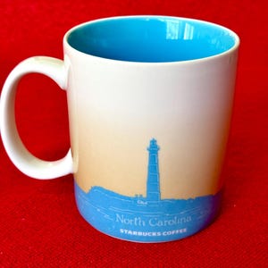 May include: White ceramic mug with a blue interior and a light blue and beige ombre design. The mug features a silhouette of a lighthouse and the text "North Carolina" and "Starbucks Coffee".