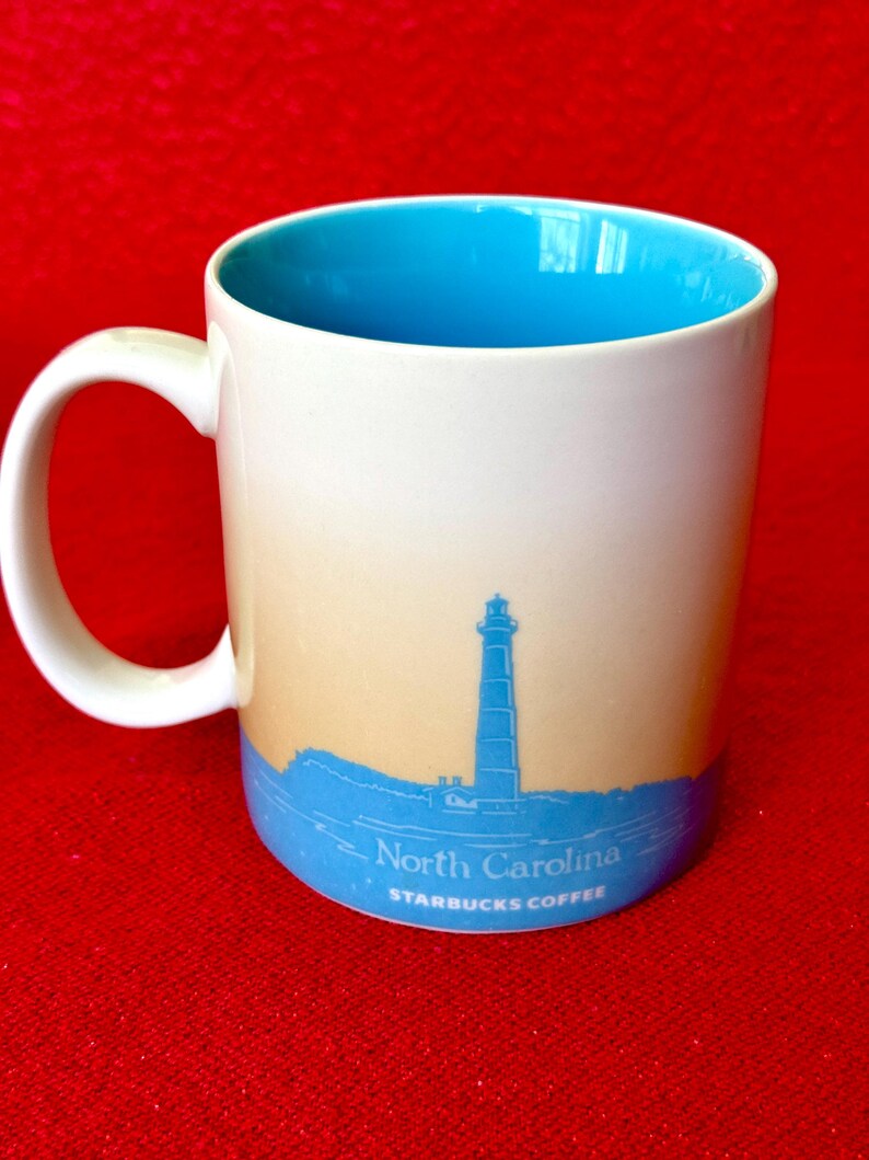 May include: White ceramic mug with a blue interior and a light blue and beige ombre design. The mug features a silhouette of a lighthouse and the text "North Carolina" and "Starbucks Coffee".