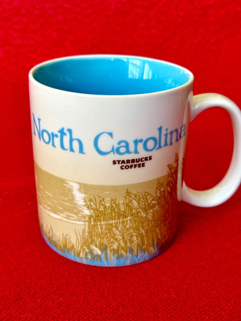 May include: White ceramic mug with a blue interior and a blue and brown image of tall grass and a beach scene. The mug has the text "North Carolina" in blue and "Starbucks Coffee" in brown.