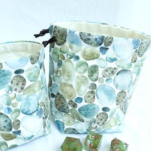 May include: Two fabric drawstring pouches featuring a pattern of blue, green, and brown river stones, paired with seven translucent green polyhedral dice with copper-colored numbers. The pouches have a cream-colored interior lining.