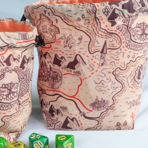 May include: Two fabric drawstring pouches featuring a beige background with a dark brown and red printed fantasy map design. The pouches have a bright orange interior lining and dark brown cord closures, surrounded by various multi-sided dice.