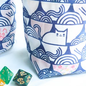 May include: Two drawstring dice bags with a white cat and blue wave pattern. The bags have a brown drawstring closure and a tan interior. Several colorful dice are in the foreground.