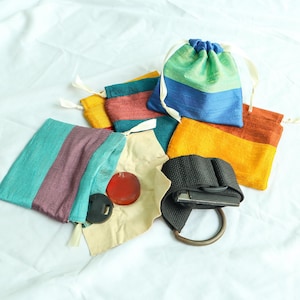 May include: An assortment of colorful drawstring bags in various sizes and hues, including blue, green, yellow, and orange. Some bags are open, revealing a red disc, a black object, and a black strap with a metal ring. The bags are made of a textured fabric.