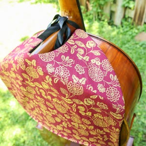 May include: A burgundy fabric cover with a gold floral pattern draped over the upper body of a wooden cello. The cover features a black ribbon tie secured around the neck of the instrument, protecting the polished wood surface from dust and wear.
