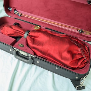 May include: A black rectangular musical instrument case with a deep red velvet interior lining. Inside, a stringed instrument is protected by a red fabric cover. A wooden bow with a floral patterned grip is secured in the lid, alongside a small gold hygrometer.