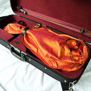 May include: A black musical instrument case with a deep red velvet interior lining. Inside, a vibrant orange and red silk drawstring bag rests in the main compartment, while a wooden bow with a floral patterned grip is secured in the lid section.