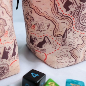 May include: Two beige fabric dice bags with a map design. The map features a red dotted line, a dragon, a church, and mountains. One bag has a black drawstring. A blue dice with the number 4 is in the foreground.