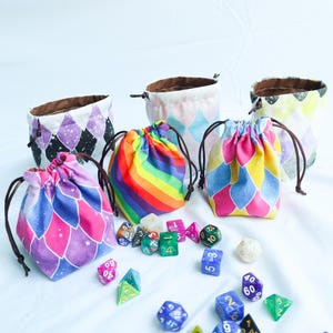 May include: A collection of colorful drawstring dice bags with various geometric patterns and a rainbow design. The bags are displayed with an assortment of polyhedral dice in different colors and sizes, perfect for tabletop gaming.