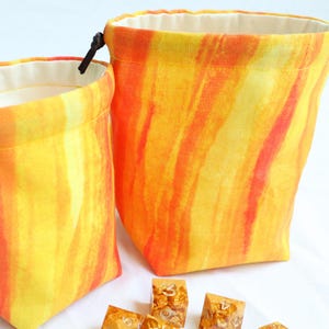 May include: Two fabric drawstring pouches featuring a vibrant yellow and orange watercolor-style vertical stripe pattern. The bags have a solid cream-colored interior lining and a dark brown cord closure. Several translucent gold-flecked dice are visible.