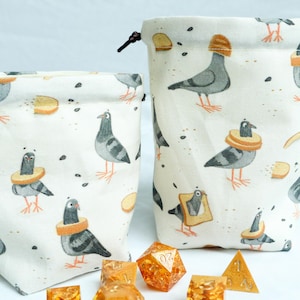 May include: Two drawstring dice bags with a repeating pattern of pigeons wearing bread hats and collars. The bags are white with a black drawstring closure. Several orange and gold dice are in front of the bags.