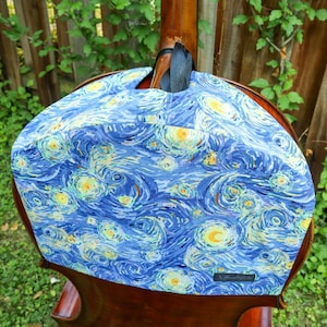 May include: A fabric cover for a stringed instrument featuring a blue and yellow swirling pattern reminiscent of a starry night sky. The cover is draped over the upper body and neck of a wooden instrument and includes a small black rectangular label.