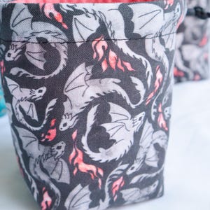 May include: A fabric dice bag with a repeating pattern of gray dragons and red flames on a dark gray background. The bag has a black drawstring closure and a pink interior. The bag is designed for tabletop gaming.