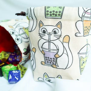 May include: Beige drawstring dice bag with a repeating pattern of cartoon cats drinking boba tea. The bag is open, revealing colorful dice inside. The bag is made of fabric.