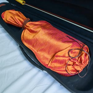 May include: An open black violin case with a vibrant orange and red silk cover. The cover is secured with a brown leather strap and drawstring. A violin bow with a yellow grip is visible.