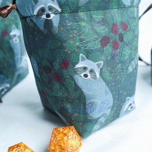 May include: A drawstring bag with a teal background featuring a pattern of raccoons, leaves, and berries. The bag is made of fabric and has a brown drawstring closure. Two orange, multi-sided dice are in the foreground.