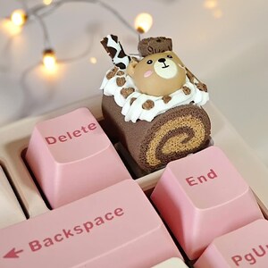 Puede incluir: Un teclado decorativo con teclas rosas y un adorno de pastel caprichoso. Las teclas incluyen "Delete", "Backspace" y "End". El adorno de pastel presenta una figura de oso sobre un pastel de rollo de chocolate con glaseado blanco y detalles de chocolate.