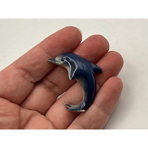 May include: A small, glossy ceramic dolphin figurine featuring a deep navy blue color on the dorsal side that transitions into a lighter, pale greyish-white tone on the underside. The dolphin is depicted in a curved, leaping posture.