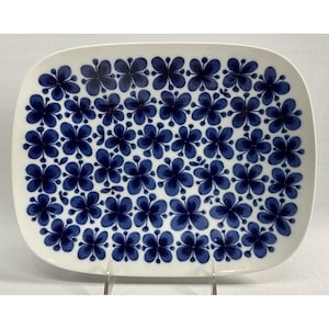 May include: A rectangular white ceramic serving platter featuring a repeating pattern of dark blue four-petalled flowers across the entire surface. The platter has rounded corners and a smooth, glossy finish with a consistent floral motif.