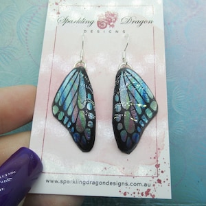 May include: Pair of iridescent blue and black butterfly wing earrings with silver hooks. The earrings are on a white card with the text "Sparkling Dragon Designs" and "www.sparklingdragondesigns.com.au".