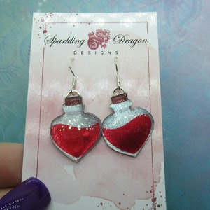 May include: A pair of silver heart-shaped earrings with red glitter potion bottles. The earrings are on a white card with the text "Sparkling Dragon Designs".