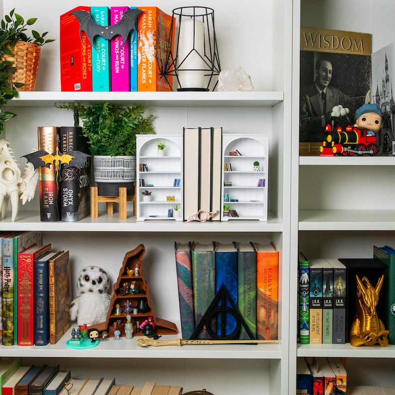 以下が含まれることがあります： 本、装飾品、植物でいっぱいの白い本棚。カラフルな背表紙の本が棚に並べられ、フィギュア、キャンドルホルダー、装飾的なコウモリが並んでいます。全体的なテーマは、文学と装飾の厳選されたコレクションです。