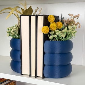 May include: Navy blue bookends with a stacked, rounded design. They hold books and are decorated with dried flowers and yellow globe-shaped blooms. The arrangement sits on a white shelf. The bookends are approximately 15 cm tall.