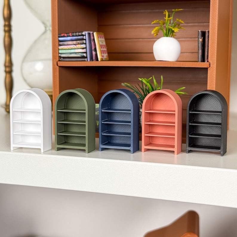 May include: Five miniature arched bookshelves in white, olive green, navy blue, coral, and black. Each shelf has three horizontal shelves. The shelves are arranged on a white surface, with a larger wooden shelf in the background.