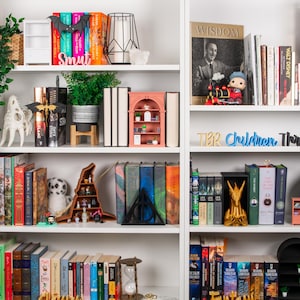 May include: A white bookcase with books, ornaments, and plants. The shelves display a variety of book spines, a miniature bookshelf, a bat-shaped decoration, and a gold-coloured statue.