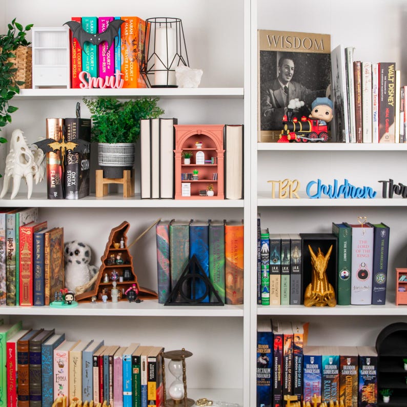 May include: A white bookcase with books, ornaments, and plants. The shelves display a variety of book spines, a miniature bookshelf, a bat-shaped decoration, and a gold-coloured statue.