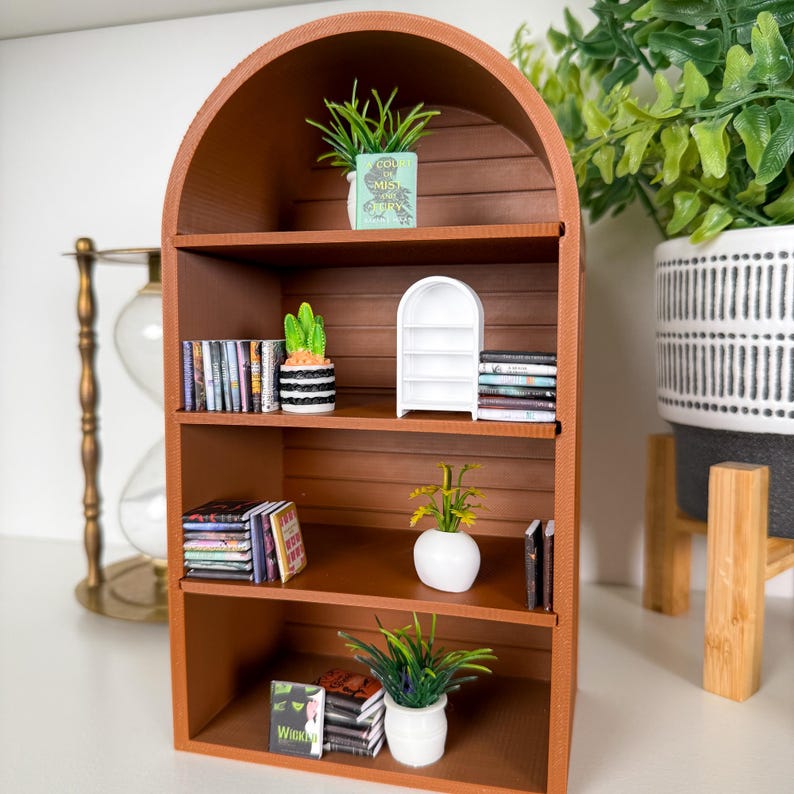 May include: A miniature brown wooden bookshelf with an arched top, filled with tiny books, potted plants, and a white decorative shelf. The plants include a cactus, a small green plant in a blue pot, and a plant with yellow flowers in a white pot.