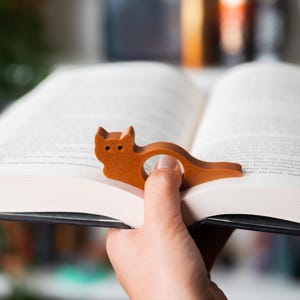 May include: A brown cat-shaped bookmark made of wood, resting on the pages of an open book. The bookmark has a circular opening for a finger to hold the book open. The cat's head and tail are visible.