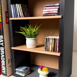 May include: A black bookshelf with three shelves. The top shelf has a stack of books with colourful covers. The middle shelf has a potted green plant and a stack of books with colourful covers. The bottom shelf has a stack of books with colourful covers and a small white planter with pink and yellow flowers.