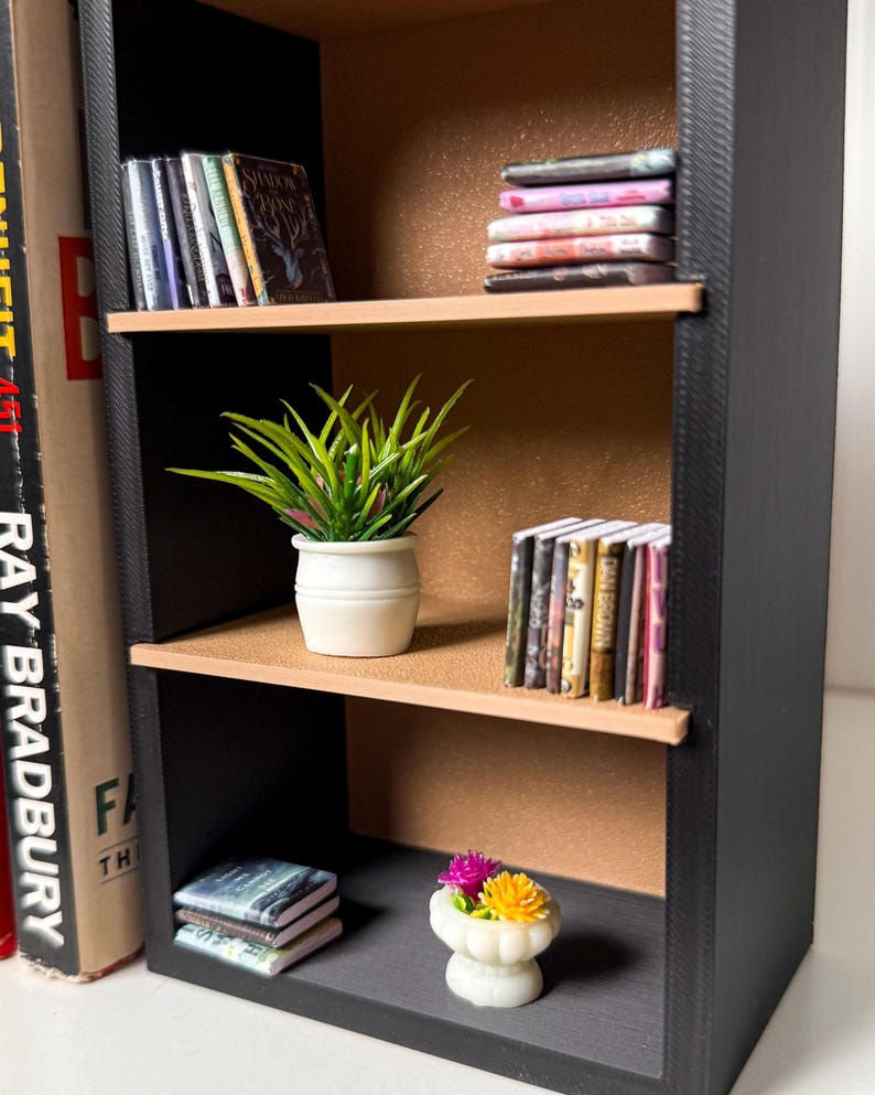 May include: A black bookshelf with three shelves. The top shelf has a stack of books with colourful covers. The middle shelf has a potted green plant and a stack of books with colourful covers. The bottom shelf has a stack of books with colourful covers and a small white planter with pink and yellow flowers.