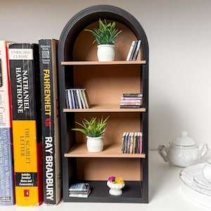 May include: A miniature black bookshelf with three shelves, featuring a selection of miniature books, potted plants, and a small flower arrangement. The bookshelf has an arched top and is made of wood.